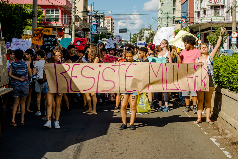 Participantes da marcha das mulheres ocupando uma avenida de Bauru carregando uma faixa com os dizeres "Resiste Mulher"