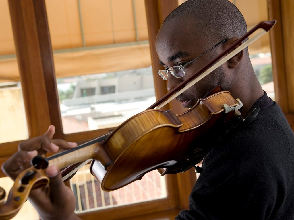 homem negro, careca, está utilizando uma camiseta preta e segurando um violino, ao fundo há uma janela para a rua