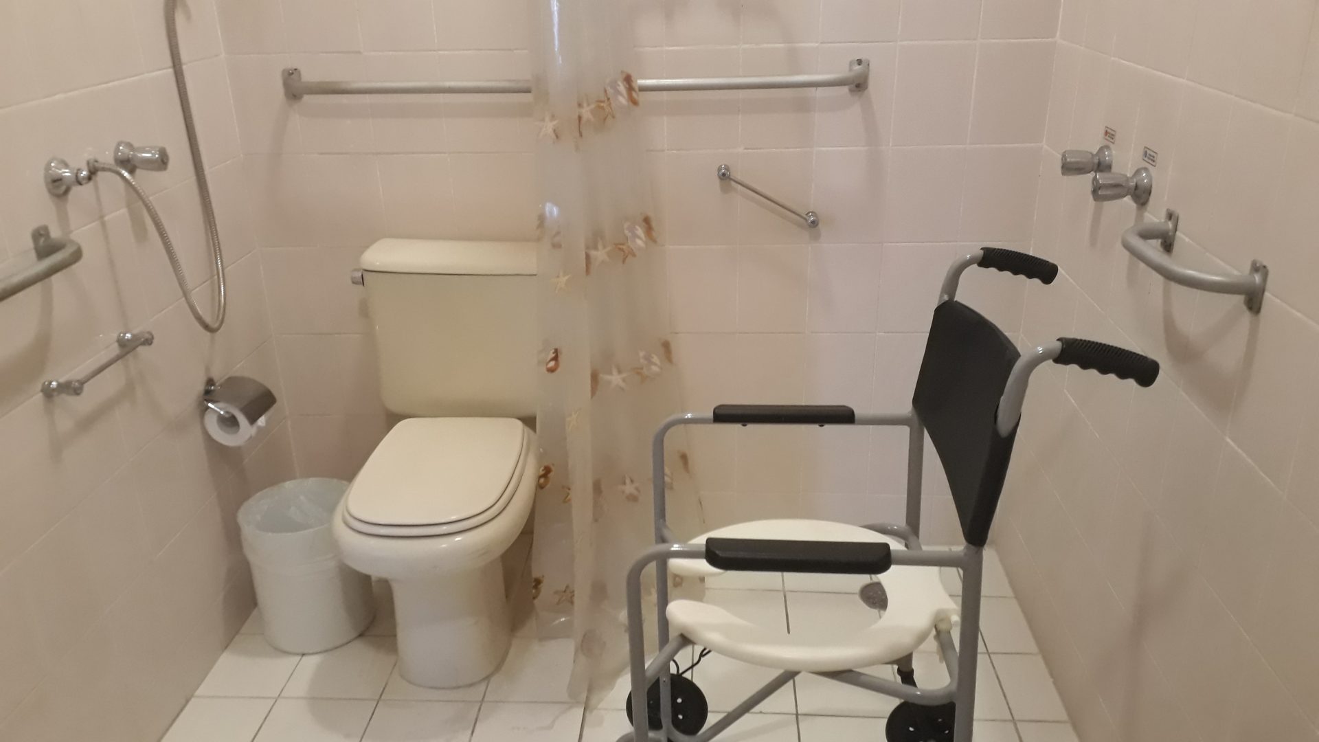 Imagem de um dos banheiros de um adaptados de um hotel. Nele se vê uma cadeira de banho para deficientes, um vaso sanitário branco e barras de apoio pregadas nas paredes. Acessibilidade