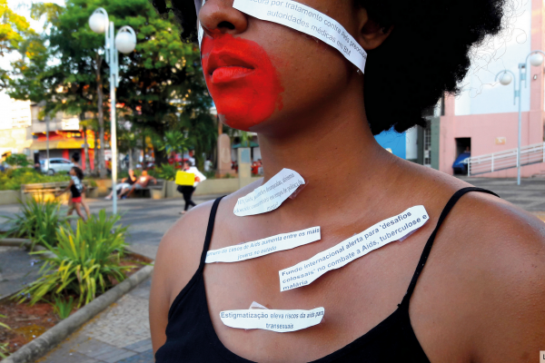 Imagem de capa. Uma mulher negra com os lábios e pele em torno dos lábios pintados de vermelho. Papéis com manchetes relacionadas ao HIV estão colados em seu corpo.