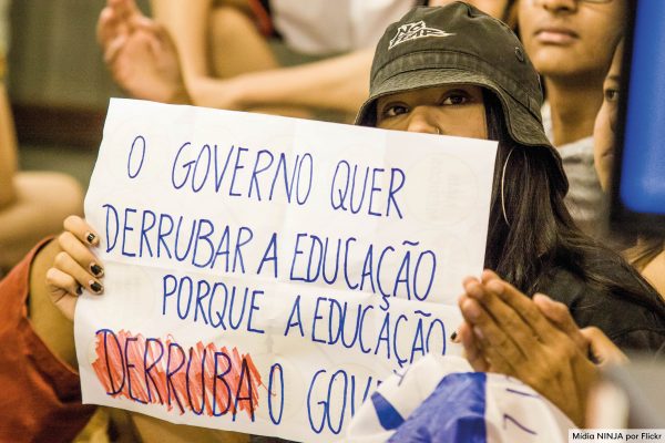 Imagem de capa. Uma moça segura um cartaz com a frase "O governo quer derrubar a educação porque a educação derruba o governo".